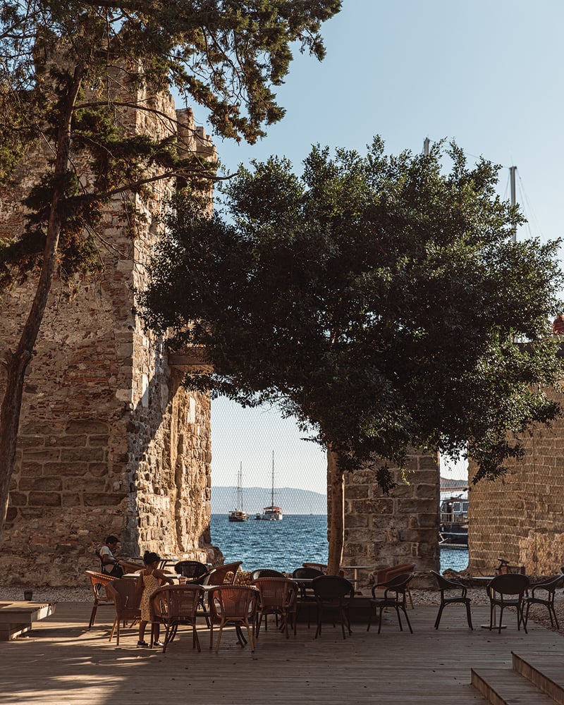 Boats in the Mediterranean Sea, seen through openings in the stone walls of medieval Bodrum Castle, with café tables and chairs in the foreground. 