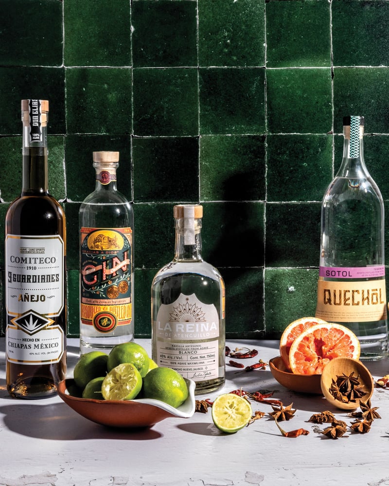 Four Mexican spirits in front of a green tile wall, with limes, chilies, grapefruit, and star anise scattered around them.