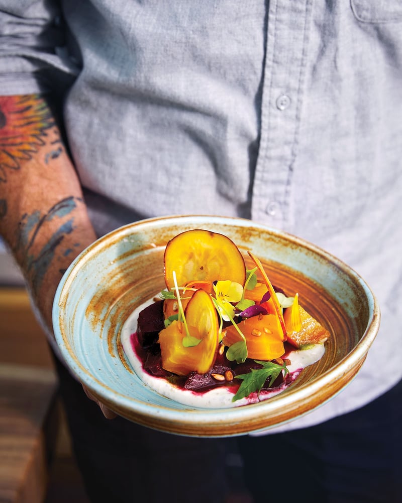 A closeup shot of a person with a light blue button-down shirt and tattooed arm holding a ceramic blue and brown bowl with fruit and vegetables in yogurt.