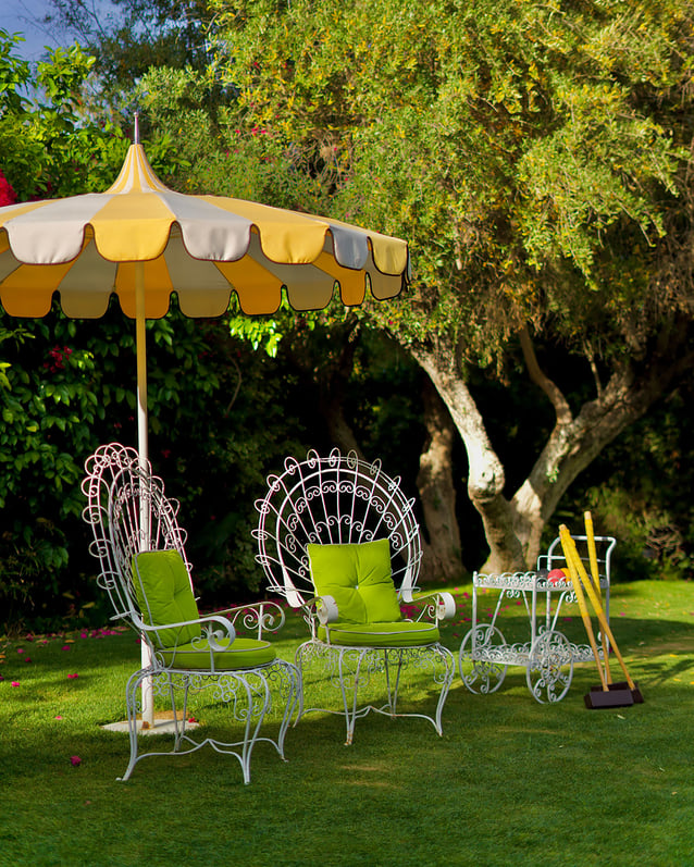 Two chairs with filigreed metal backs and green cushions under a yellow-and-white-striped umbrella on the croquet lawn at The Parker Palm Springs.