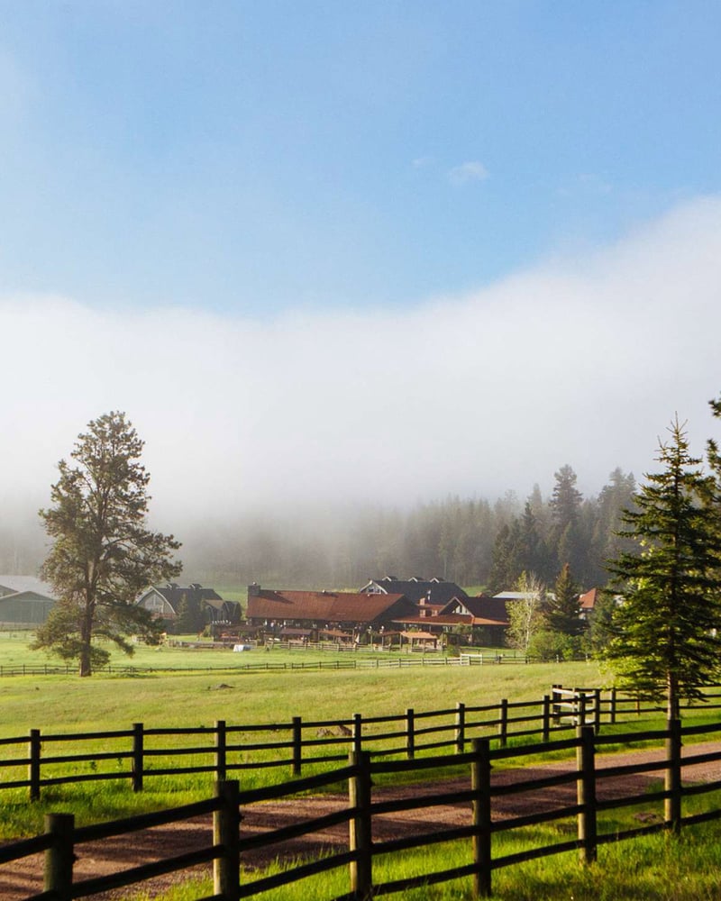 A cabin in a green field, near a grove of trees and a red-dirt road lined by a wooden fence, at Montana’s Resort at Paws Up.