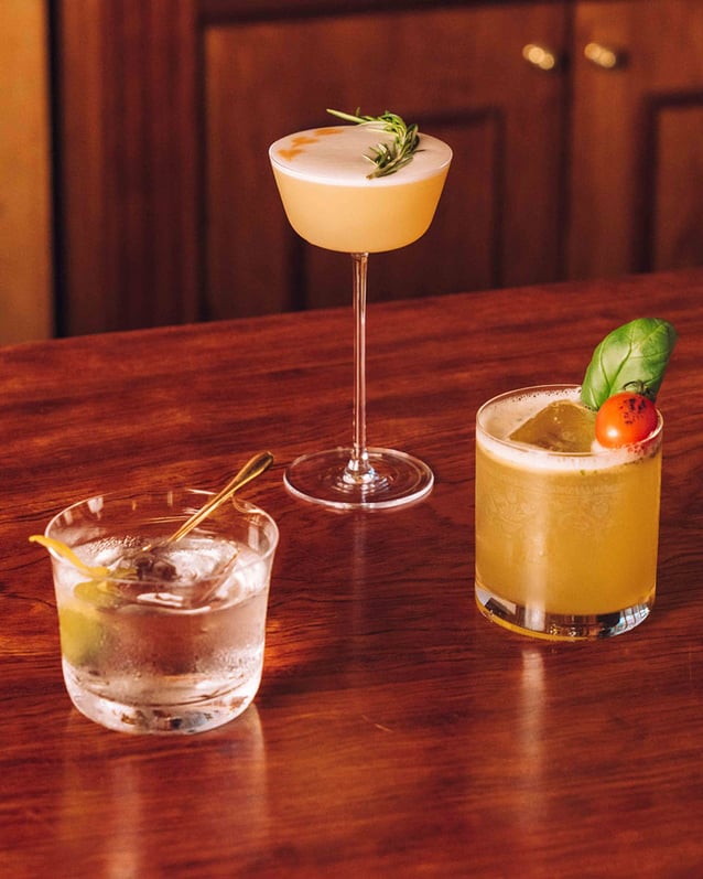 Three cocktails, two in lowball glasses and one in a coupe, rest on a wooden bar at Cobbler.