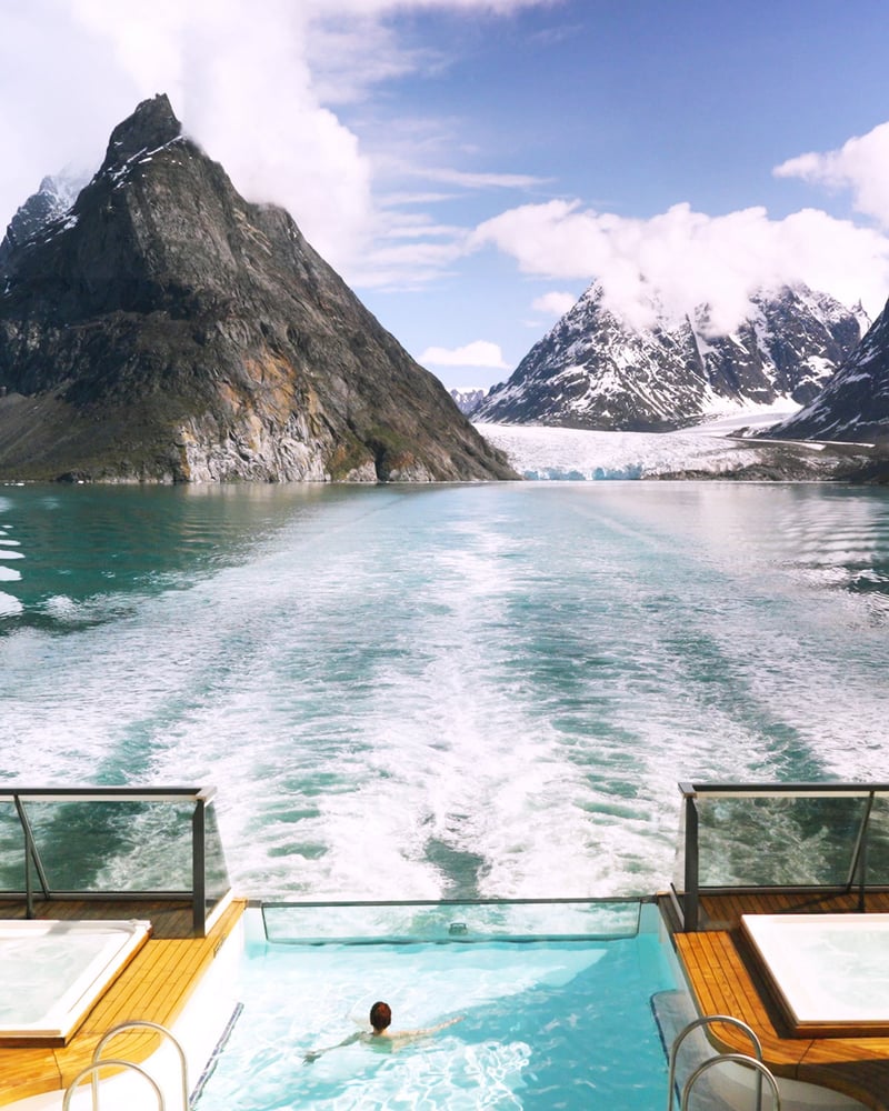 A woman looks out at Greenland’s mountains from an outdoor pool on a ship deck.