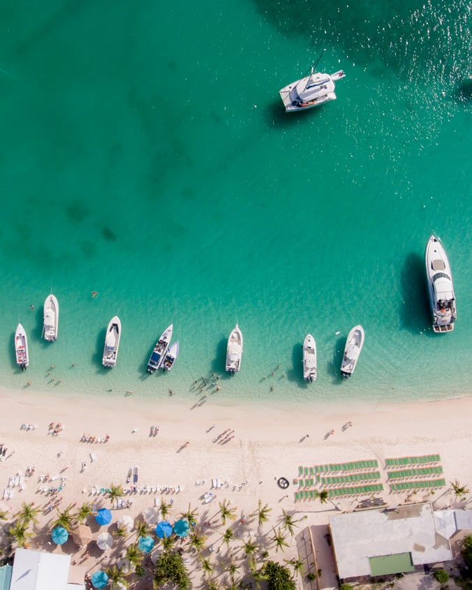 An overhead view of White Bay beach in the BVI, with boats anchored at the shore and people lining the beach.