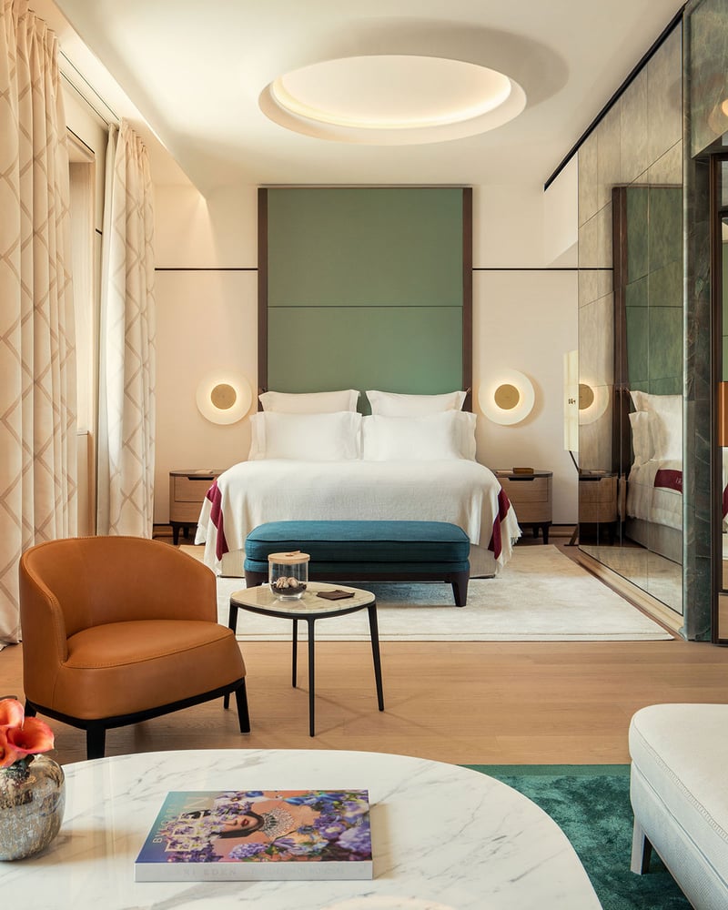 A Bulgari Hotel Roma guest room with marigold and teal furniture, an emerald rug, a white bed, and a sage accent wall.