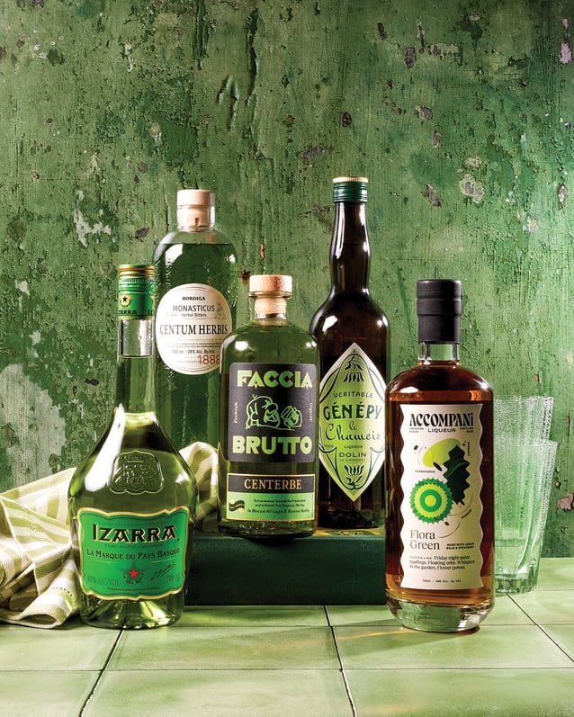 Five bottles of Chartreuse alternatives, drinking glasses, and a green and white striped napkin against a green background.