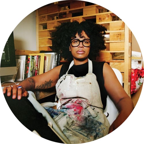 Allison Janae Hamilton, wearing chunky glasses, a black shirt, and an apron, gazes at the camera from inside her art studio.
