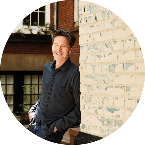 Andrew McCarthy leans against a white brick wall and smiles at the camera.