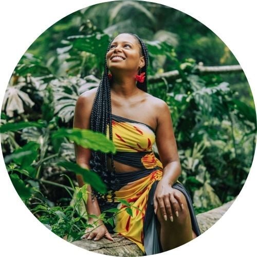 Brittany Johnson, wearing a floral-print dress and red earrings and sitting among greenery, looks upward and smiles.