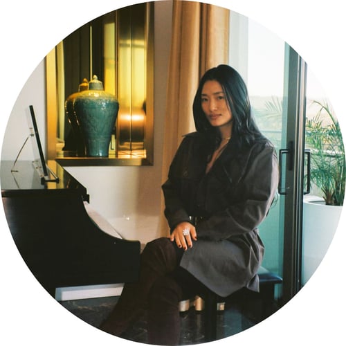 Chloe Flower, wearing an oversize blazer and sitting at a piano, smiles softly at the camera.