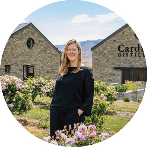 Desiree Reid, wearing black, stands among pink roses on the lawn of Cardrona Distillery and smiles at the camera.
