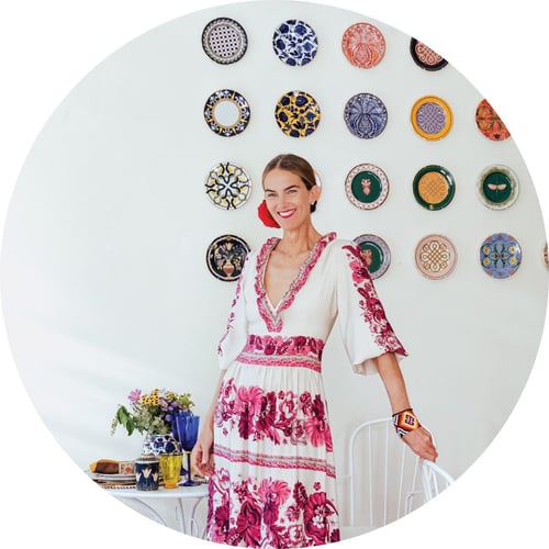 J.J. Martin, wearing a V-necked, pink-and-white maxi dress, smiles at the camera. She stands in front of a wall decorated with colorful plates.