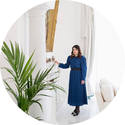 Wearing a blue dress, Mariella Avino stands in a white room with potted plants in the foreground.