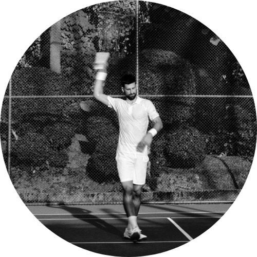Novak Djokovic, wearing all white, demonstrates a move on the tennis court in a black-and-white photo.