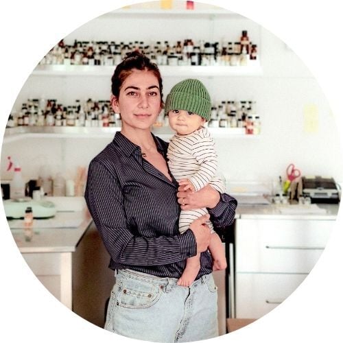 Shabnam Tavakol, wearing a striped button-down and holding her young daughter, stands in front of a wall of fragrances and smiles at the camera.