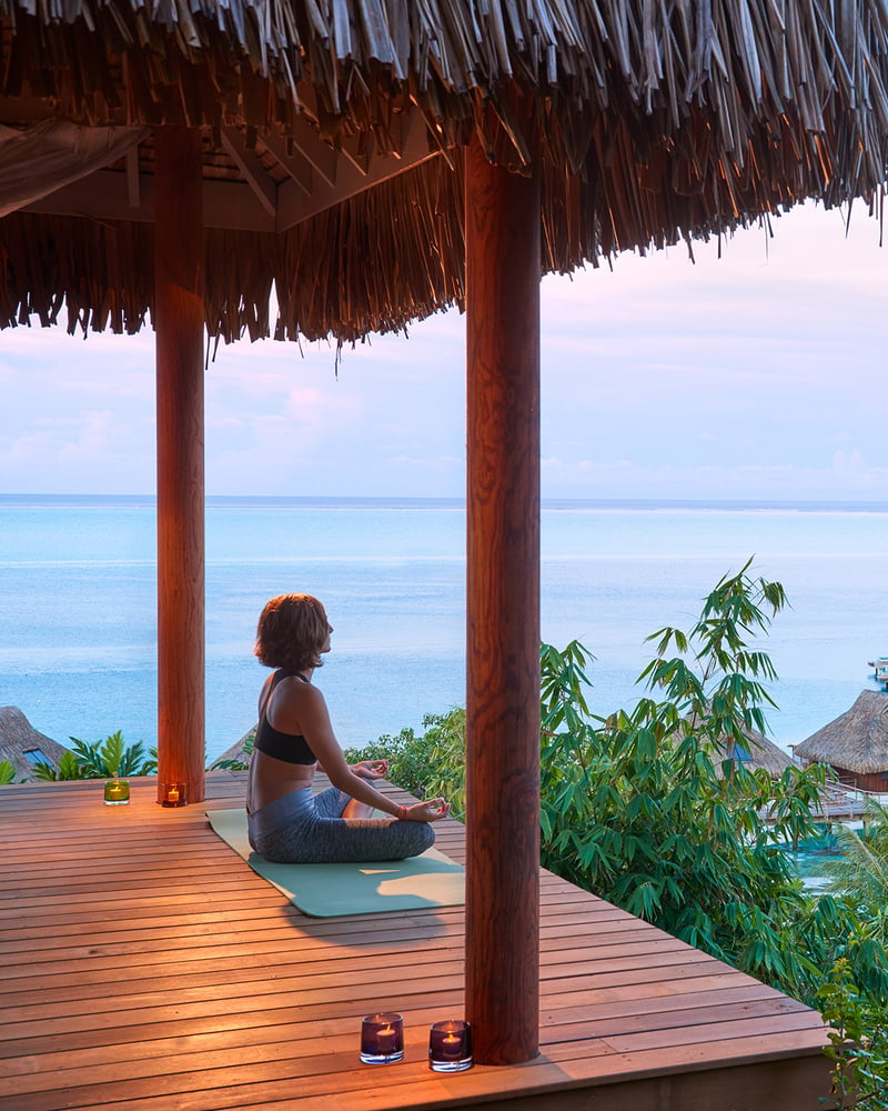 A woman meditates in an open thatched pavilion with a sunset view over the ocean at Conrad Bora Bora Nui.