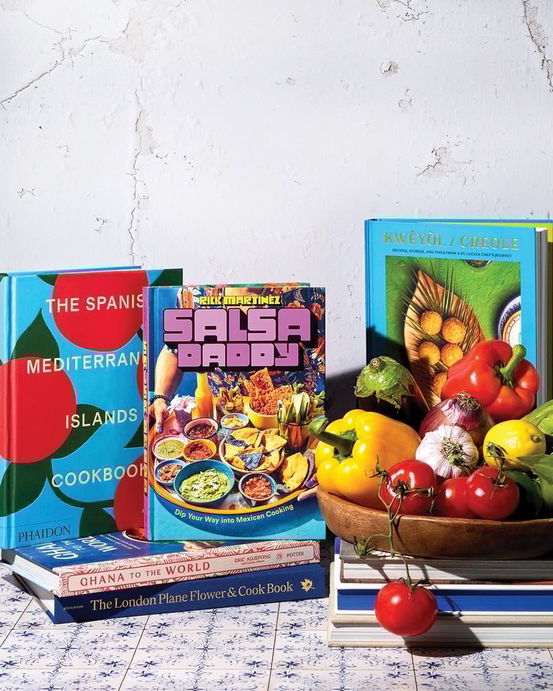 Five colorful cookbooks are stacked on a tiled table near a bowl of veggies.