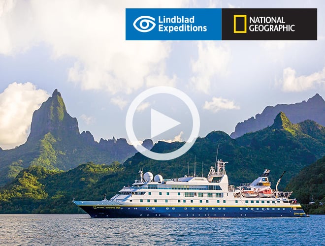Photo of a Lindblad Expeditions-National Geographic ship at sea with green mountains in the background