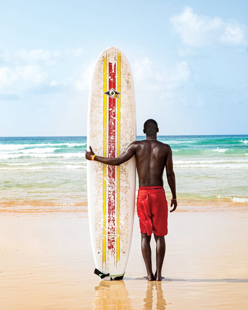 A surfer stands holding his surfboard next to him, looking out at the waves in Dakar, Senegal.