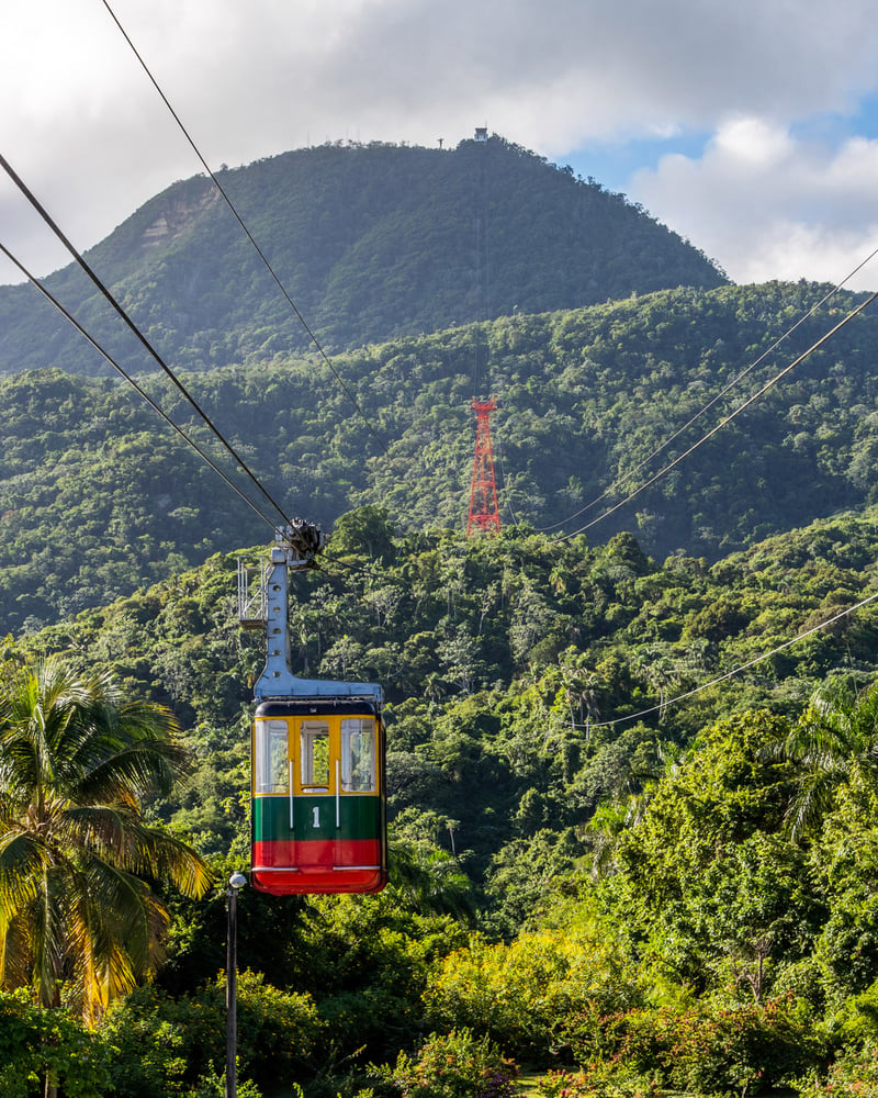 A red, yellow, and green cable car rises above the hills of the Dominican Republic.