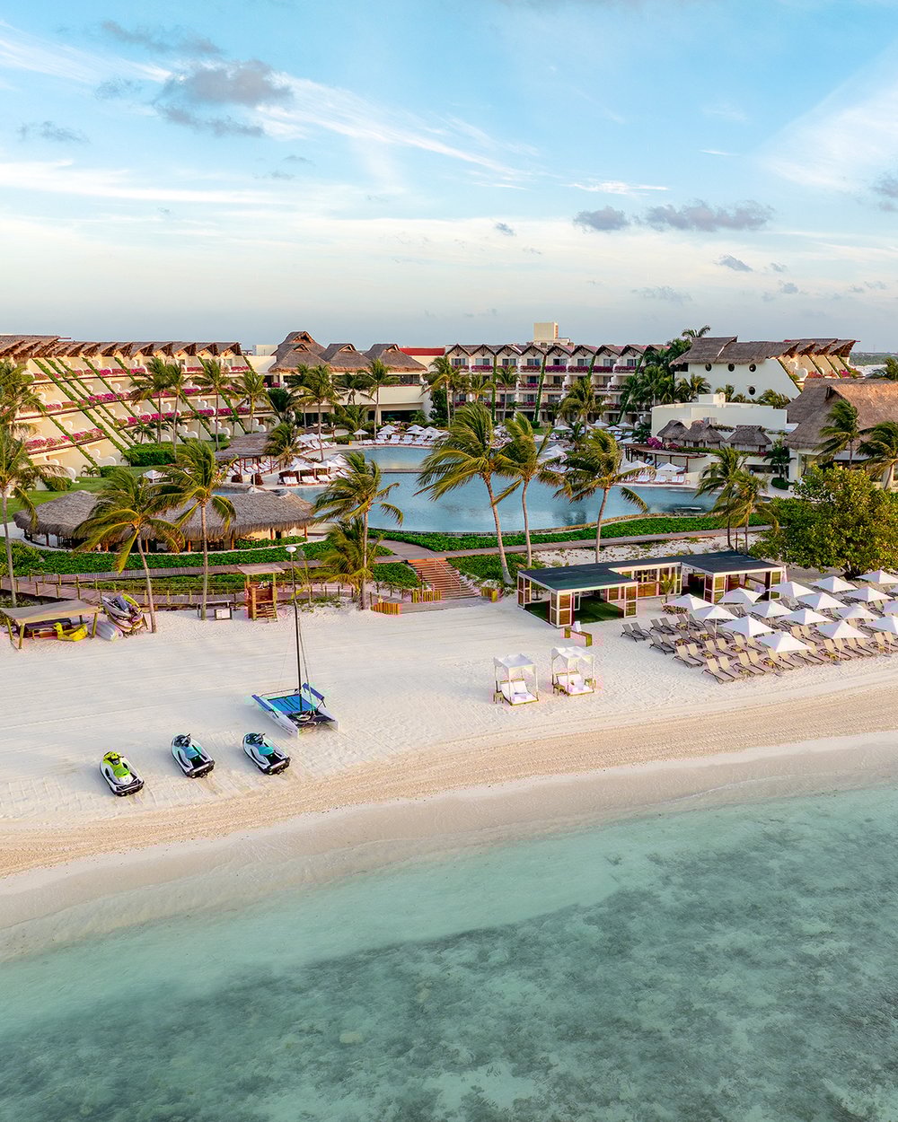 An aerial view of Grand Velas Riviera Maya’s white-sand beach, turquoise waters, palm-lined shore, and resort buildings.