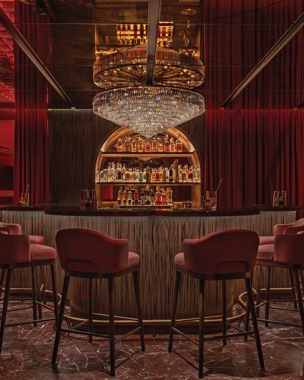 Delano Dubai’s Rose Bar, with a large crystal chandelier, red bar stools, and an oxblood-colored marble floor, all surrounded by red velvet curtains.