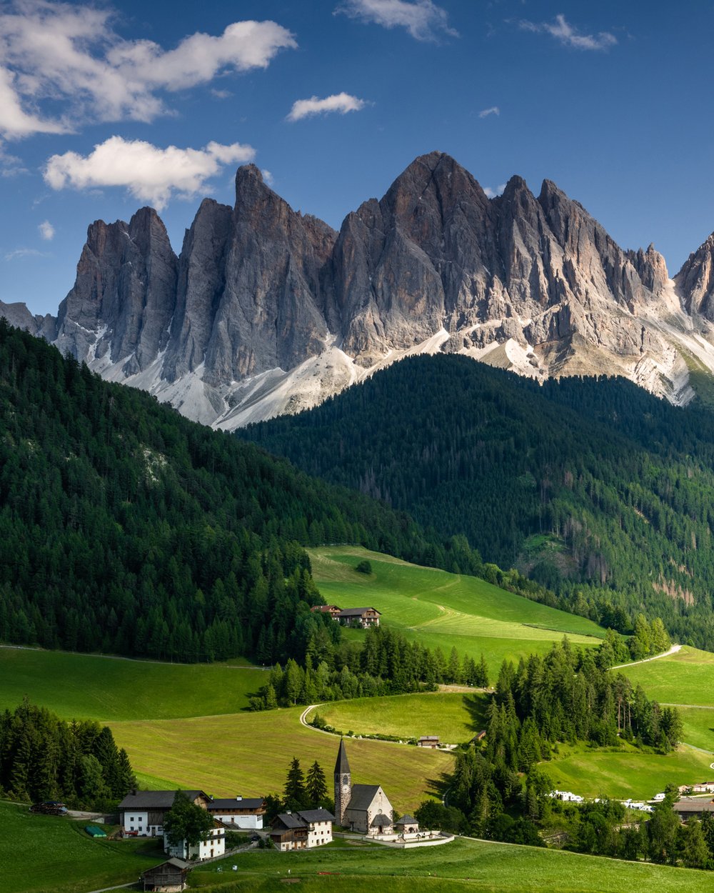 Small houses sit on a grassy field with the rocky Dolomites rising in the distance.