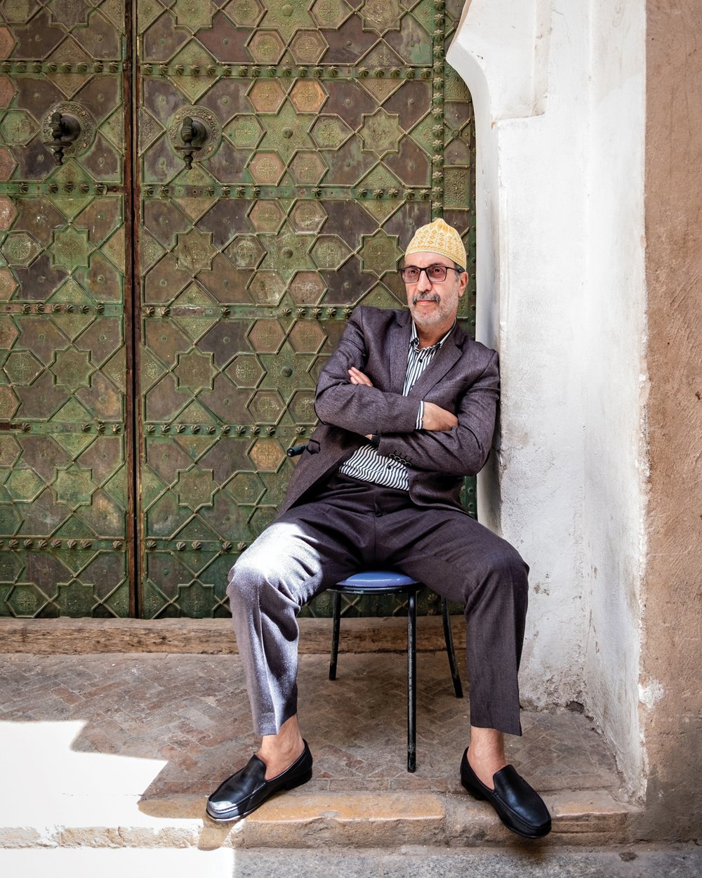 A man sits at ease on a stool in front of a verdigris engraved door at a Fez medina.