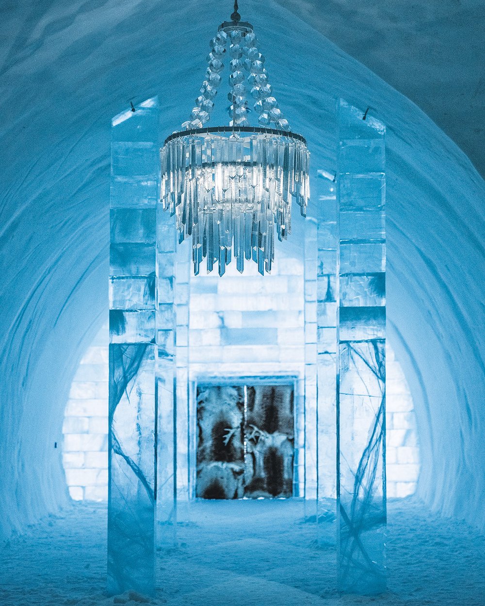 A corridor with a chandelier leads to a door, all made of ice.