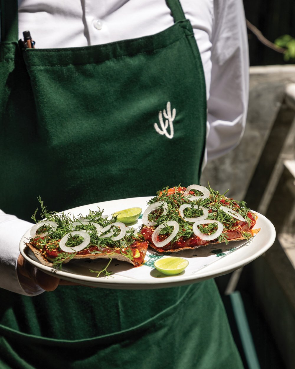 A server wearing a green apron holds a plate with two tuna tostadas, garnished with greens, onions, and limes.