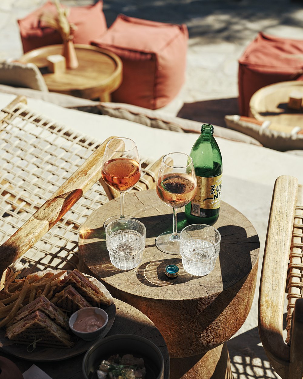 A rustic wooden table between two caned wood chairs holds two glasses of wine and seltzer at the outdoor Monastiri Beach Bar.