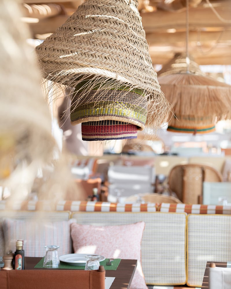 Rattan lamps hang above pale-pink, orange, and yellow textiles from design house Loro Piana at a table at La Réserve Ramatuelle’s beach club.