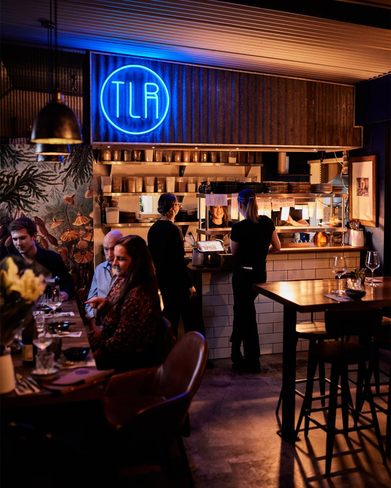 People sit in a dark bar at night eating and drinking with a neon sign in the background.