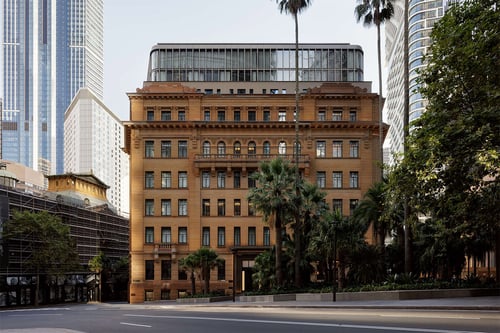 An historic hotel building stands between skyscrapers on a tree-lined street. 