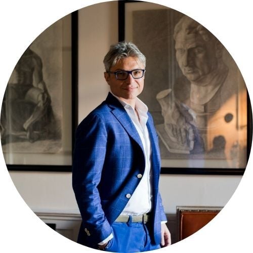 Claudio Meli, wearing a blue suit and standing in front of framed sketches, smiles at the camera.