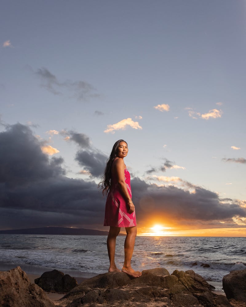 Outrigger navigator Pa’anaakala Baybayan Tanaka stands on a rock facing the ocean at sunset.