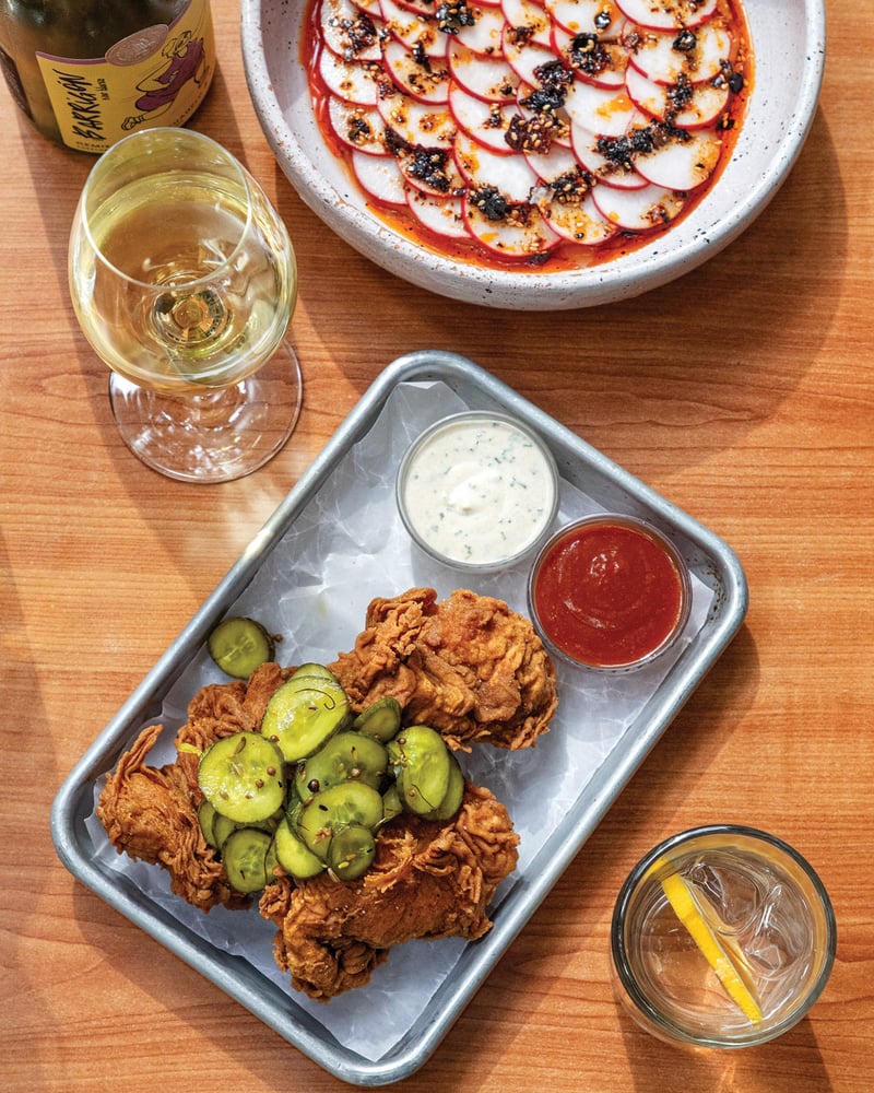 Seen from overhead is a quarter-sheet pan with fried chicken and pickles, next to a bowl of sliced radishes and some drinks.