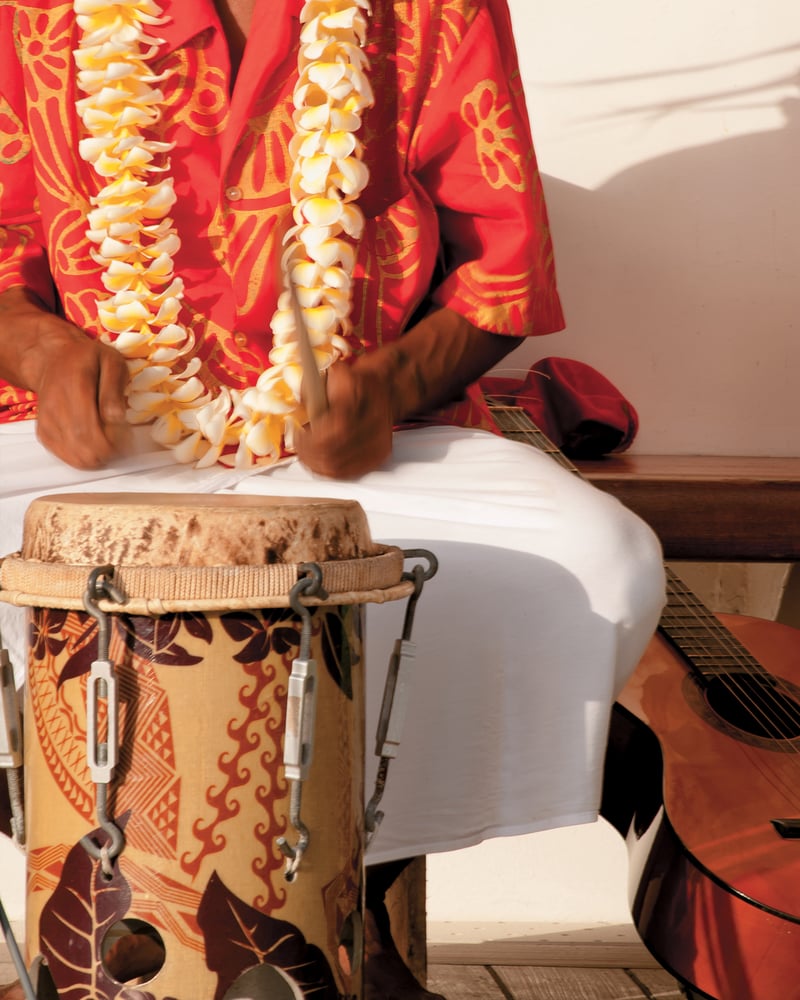 A person wearing a red printed shirt and a lei bangs on a traditional French Polynesian drum.