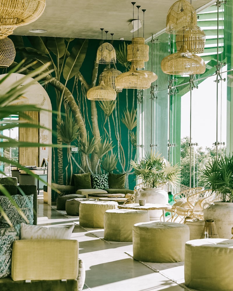 Palmaïa’s Plantissa café, with furniture in green and brown tones, hand-painted botanical wallpaper, bamboo fixtures, and full-wall windows.