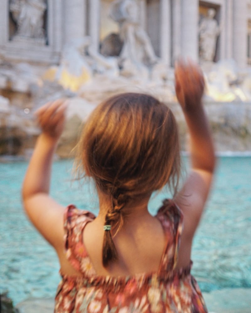 A young girl, seen from behind, tosses a coin into Rome’s Trevi Fountain.