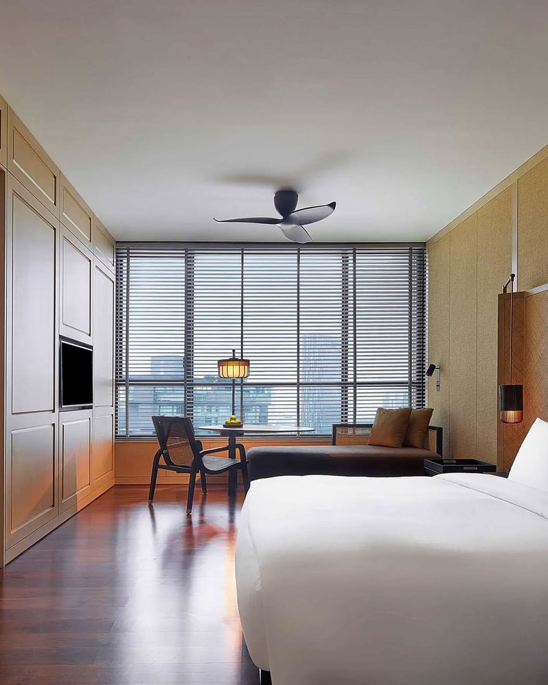 A bed and rattan-and-wood furniture in a hotel room at The RuMa, with a window overlooking Kuala Lumpur.