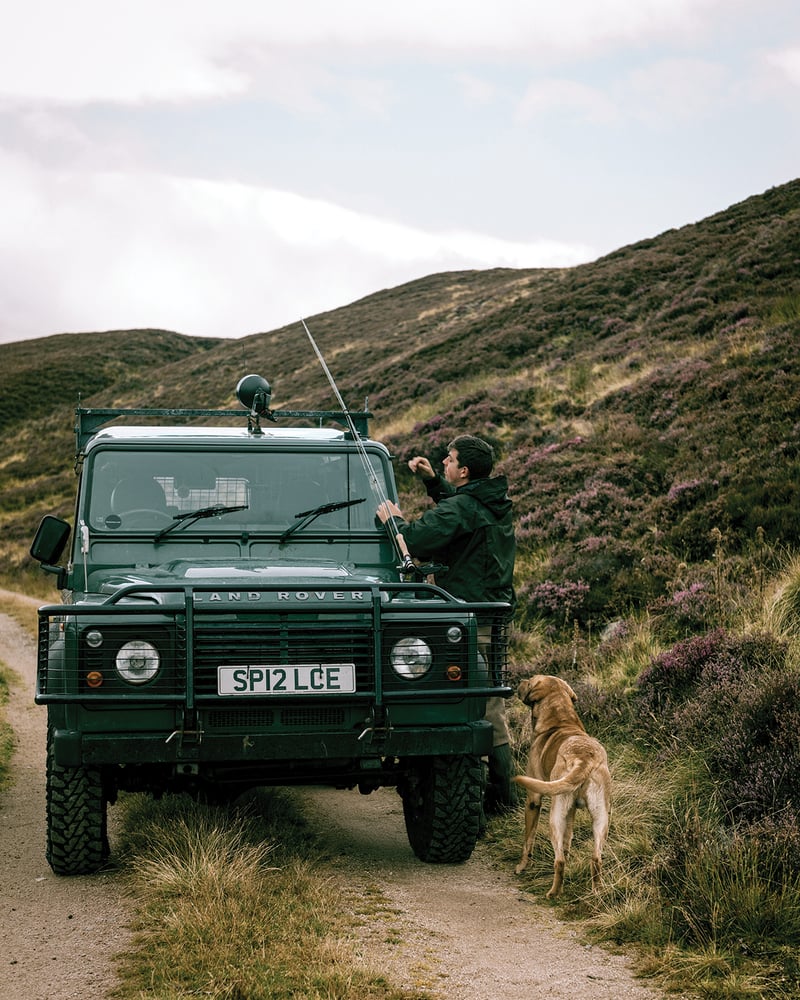 A guide unloads fishing gear from a green Range Rover in the Scottish Highlands, while a Labrador watches.