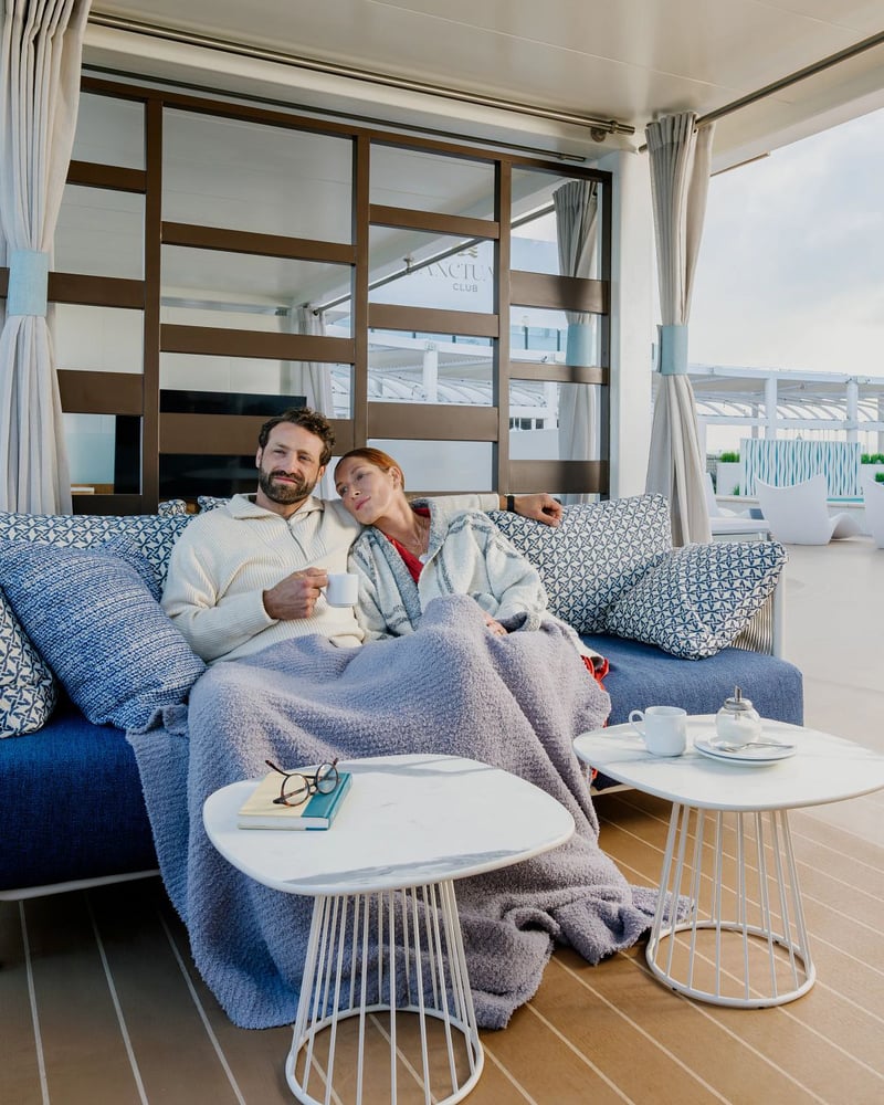 A couple relaxes on a cozy outdoor sofa under a blanket in Princess Cruises' Sanctuary Club.