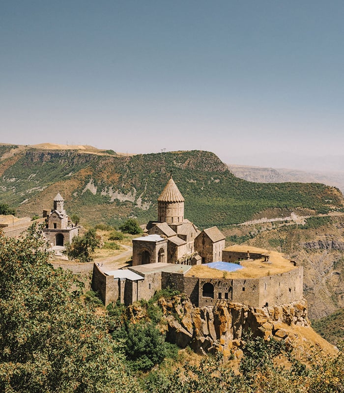 A stone monastery at the edge of a cliff, surrounded by leafy trees.