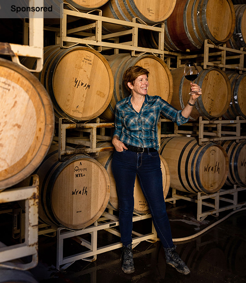 A woman wearing a flannel shirt and jeans stands before a wall of wine casks.