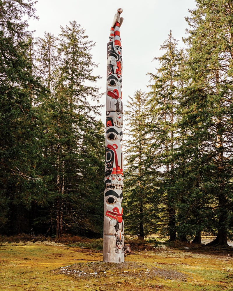 A painted totem pole rises from the ground, surrounded by trees.