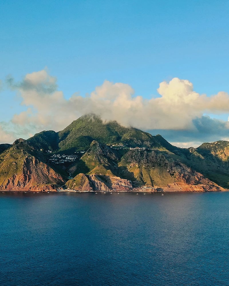 A small island with brown cliffs and green peaks stands in the blue sea on a sunny day. 