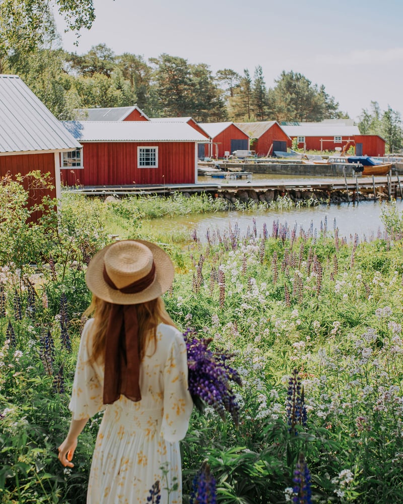 A woman with blonde hair, seen from behind, stands in a flower field facing small red cabins on the Finnish coast.