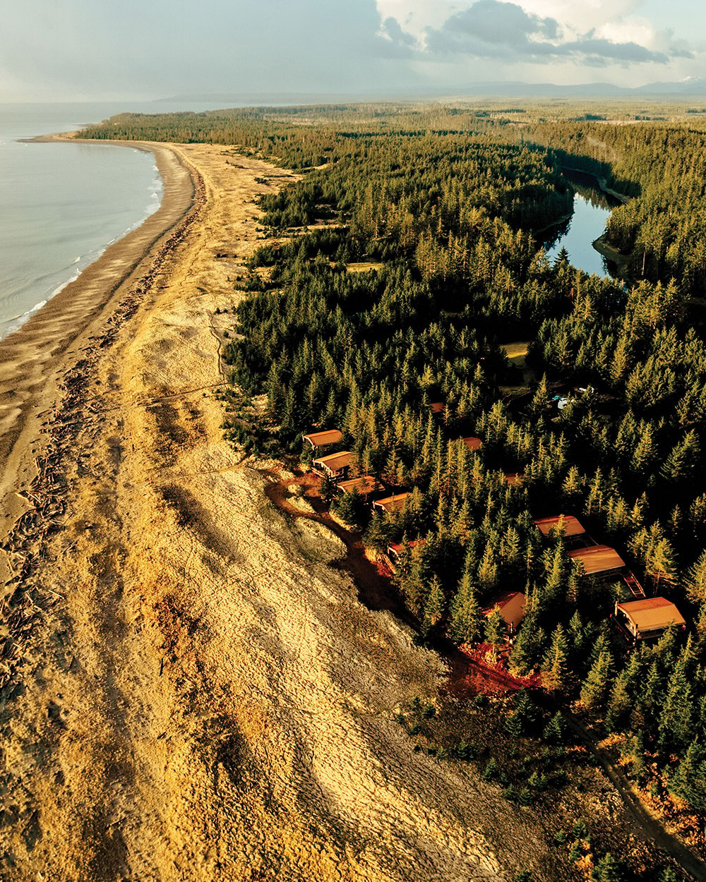 An aerial shot shows cabins in a forested area of Haida Gwaii, British Columbia, bordered to the left by golden sands and ocean.