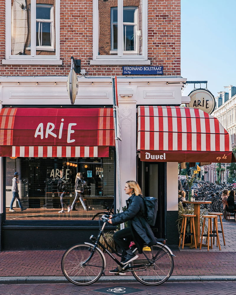 A cyclist rolls past Arie, an Amsterdam café with a red-and-white-striped awning.